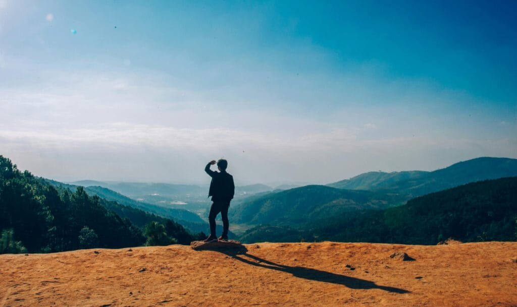 A lone traveler stands on a hilltop, gazing over a vast mountain landscape under a clear blue sky.